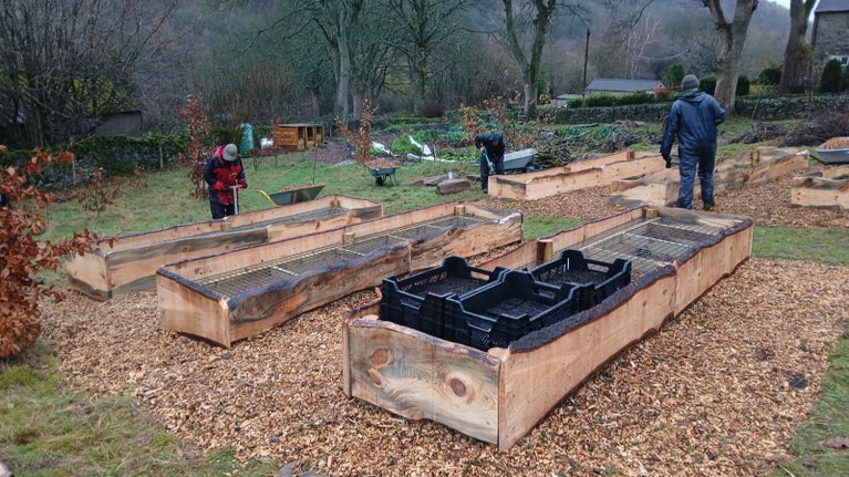 Three empty wooden raised beds in the foreground and 3 others in the distance in a large garden area with three people doing various jobs to create the growing area